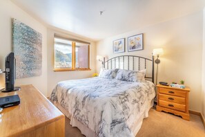 A bedroom with a bed, a wooden desk, a TV, a window with a view, and a nightstand with a lamp.
