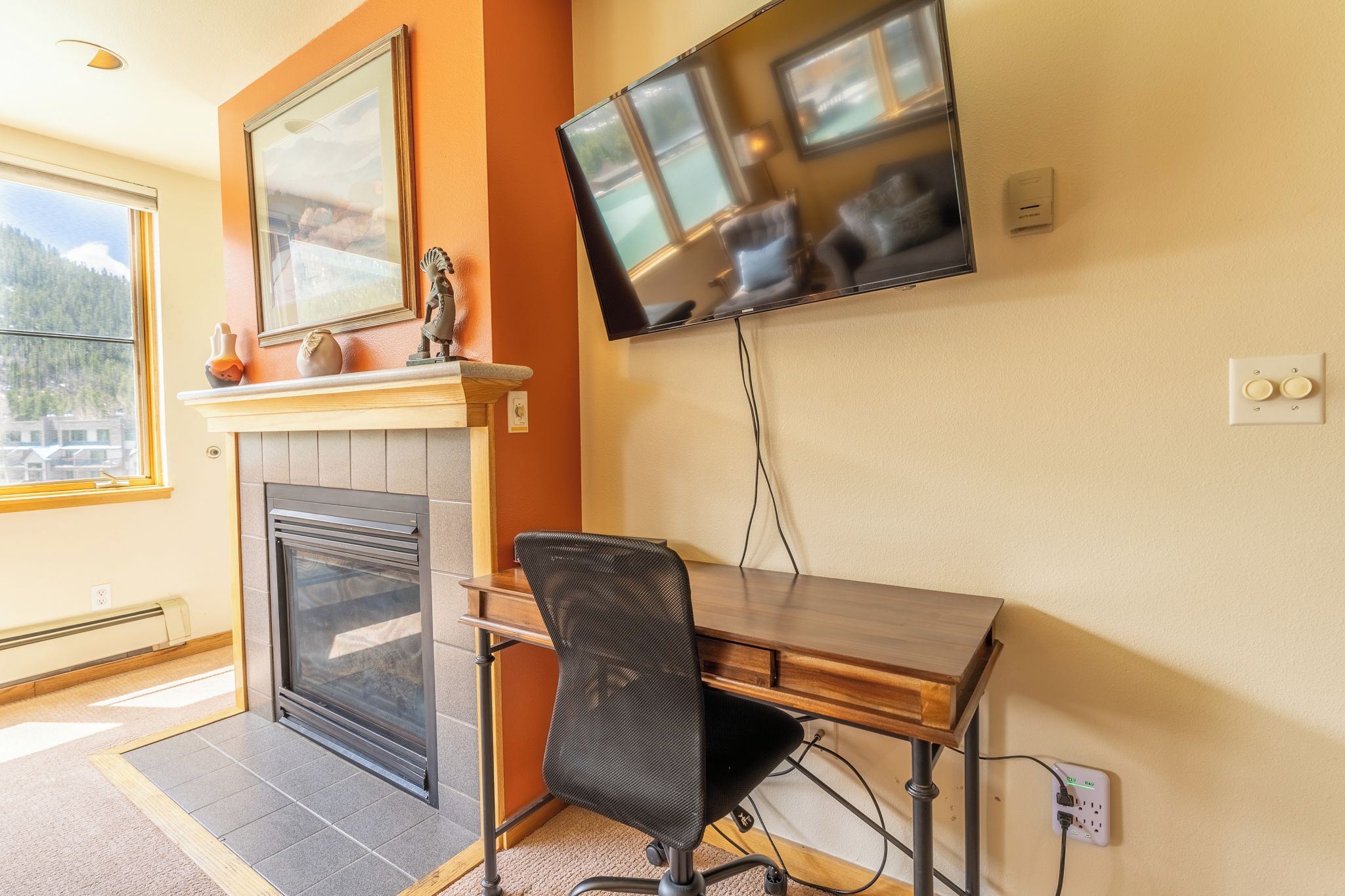 Quarto com lareira, escrivaninha com cadeira, TV fixada na parede e janela com vista para uma montanha.
