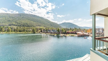 A lakeside view from a balcony with a wooden deck, overlooking a calm lake, a forested mountain, and a small town with buildings and a dock.