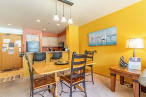 Un coin repas avec une table et des chaises en bois, une cuisine en arrière-plan et un mur jaune orné d’un tableau.