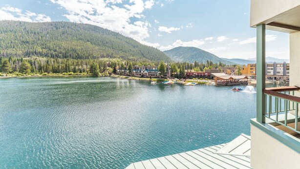 A lakeside view from a balcony with a wooden deck, overlooking a calm lake, a forested mountain, and a small town with buildings and a dock. - La1540-1 (Keystone)