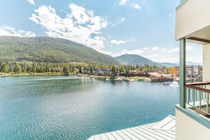 A lakeside view from a balcony with a wooden deck, overlooking a calm lake, a forested hillside, and a small town with buildings and boats.
