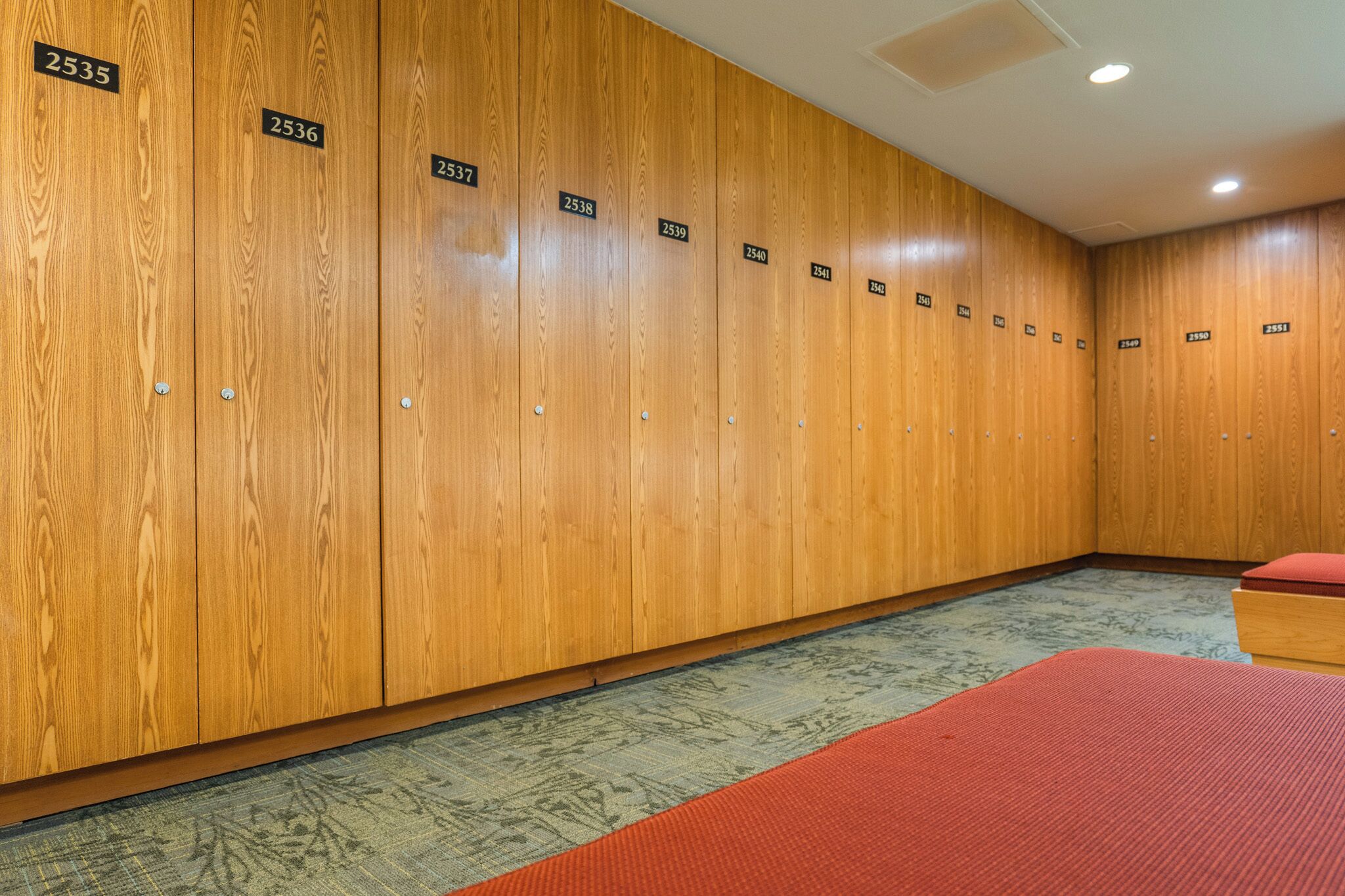 A row of wooden lockers with numbered tags, a patterned carpet, and a red bench.