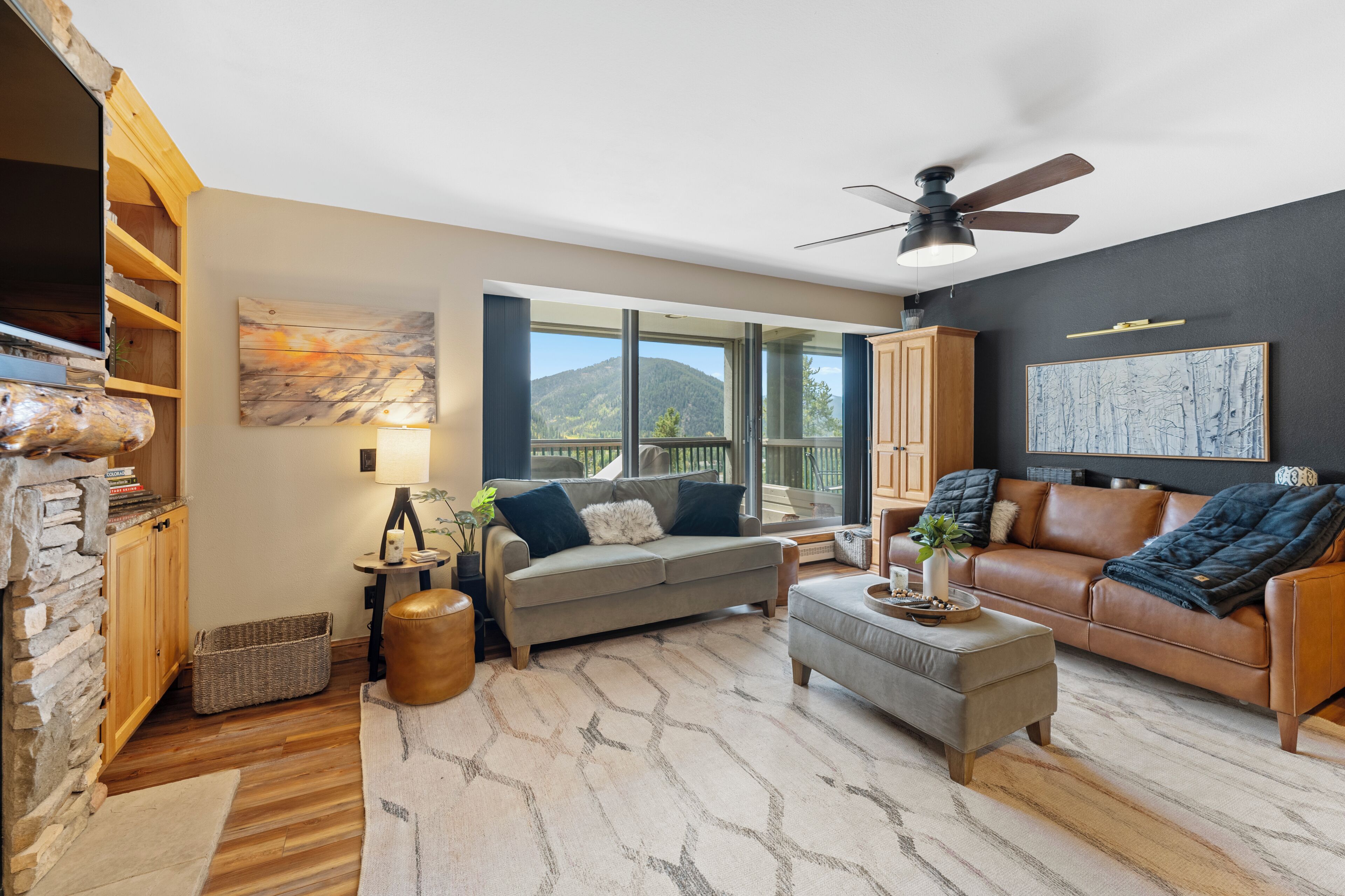 A modern living room with a stone fireplace, a large window offering a mountain view, a ceiling fan, and a cozy seating area with a leather couch and ottoman.