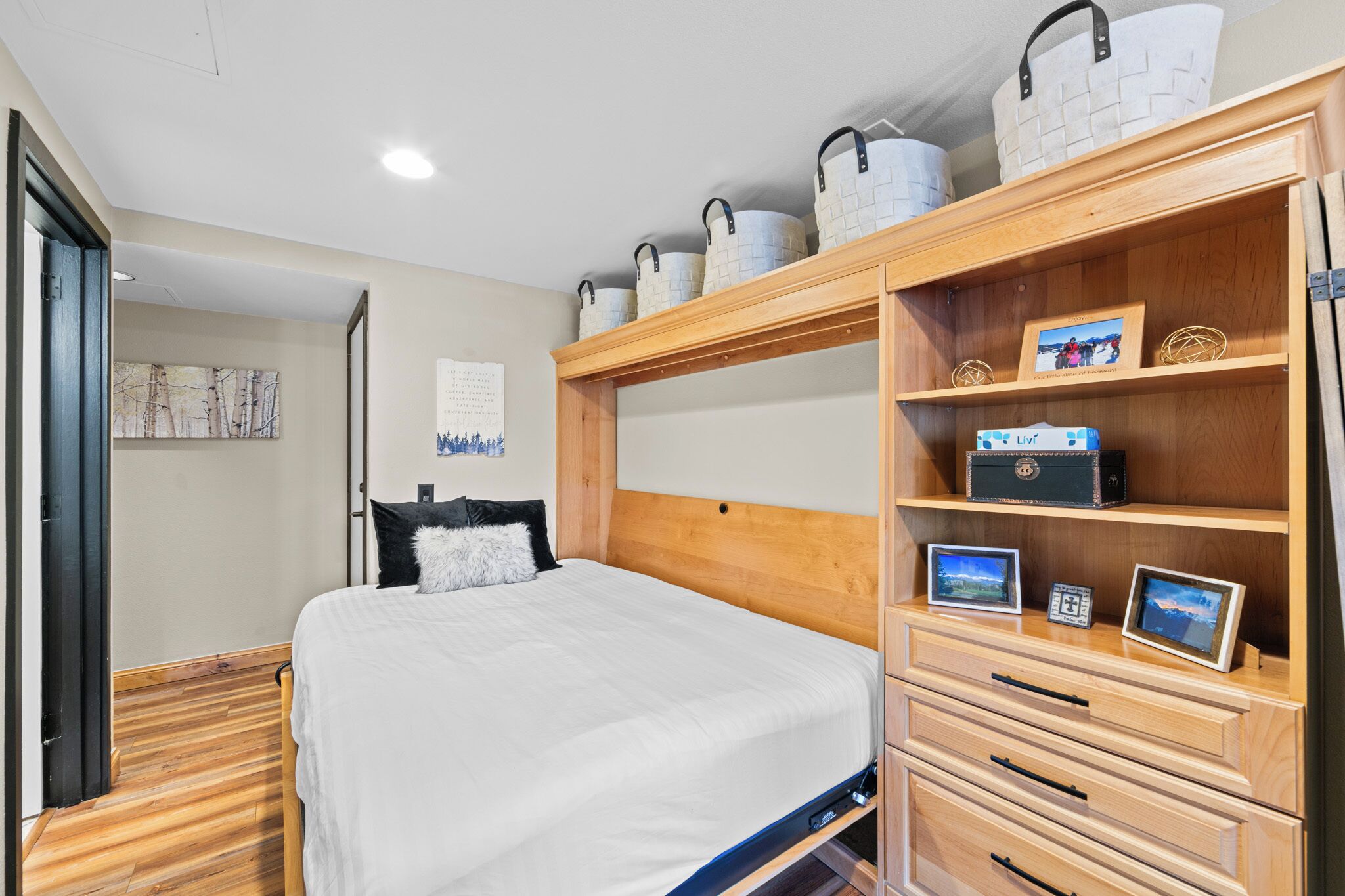 Een slaapkamer met een houten hoofdbord, een bed met witte beddengoed, een houten dressoir met lades, en een plank met decoratieve objecten en ingelijste foto’s.