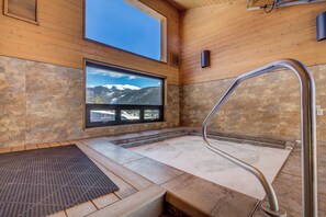 Ein Wellnessbereich mit Steinboden, einer Holzbank, einem Fenster mit Bergblick und einer Metallhandläufe.