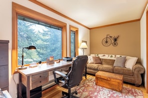 A cozy room with a wooden desk, a leather chair, a sofa, and a large window offering a view of trees.