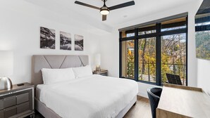 Une chambre moderne avec un grand lit, un ventilateur de plafond et une vue sur les arbres d’automne, visible à travers une grande fenêtre.