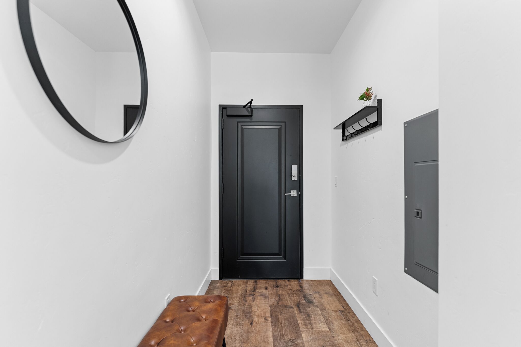 A hallway with a black door, a round mirror, a bench, and a shelf with a plant.