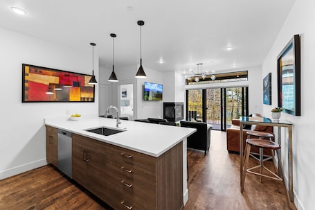 A modern kitchen with a large island, wooden cabinets, and pendant lighting. There is a dining area with a bar stool and a television.