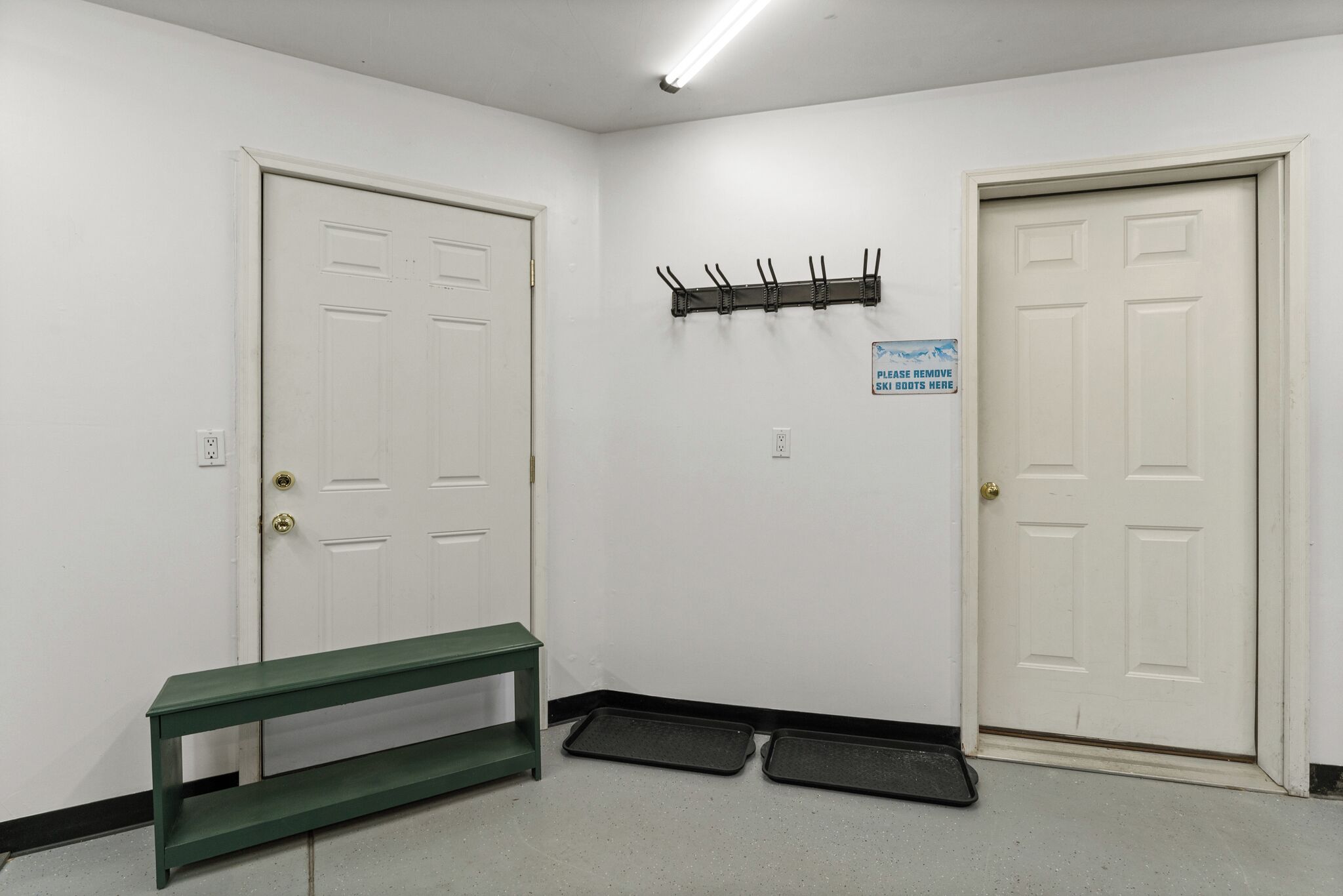 Un couloir avec deux portes blanches, un banc vert et un panneau demandant aux personnes de retirer leurs chaussures de ski.