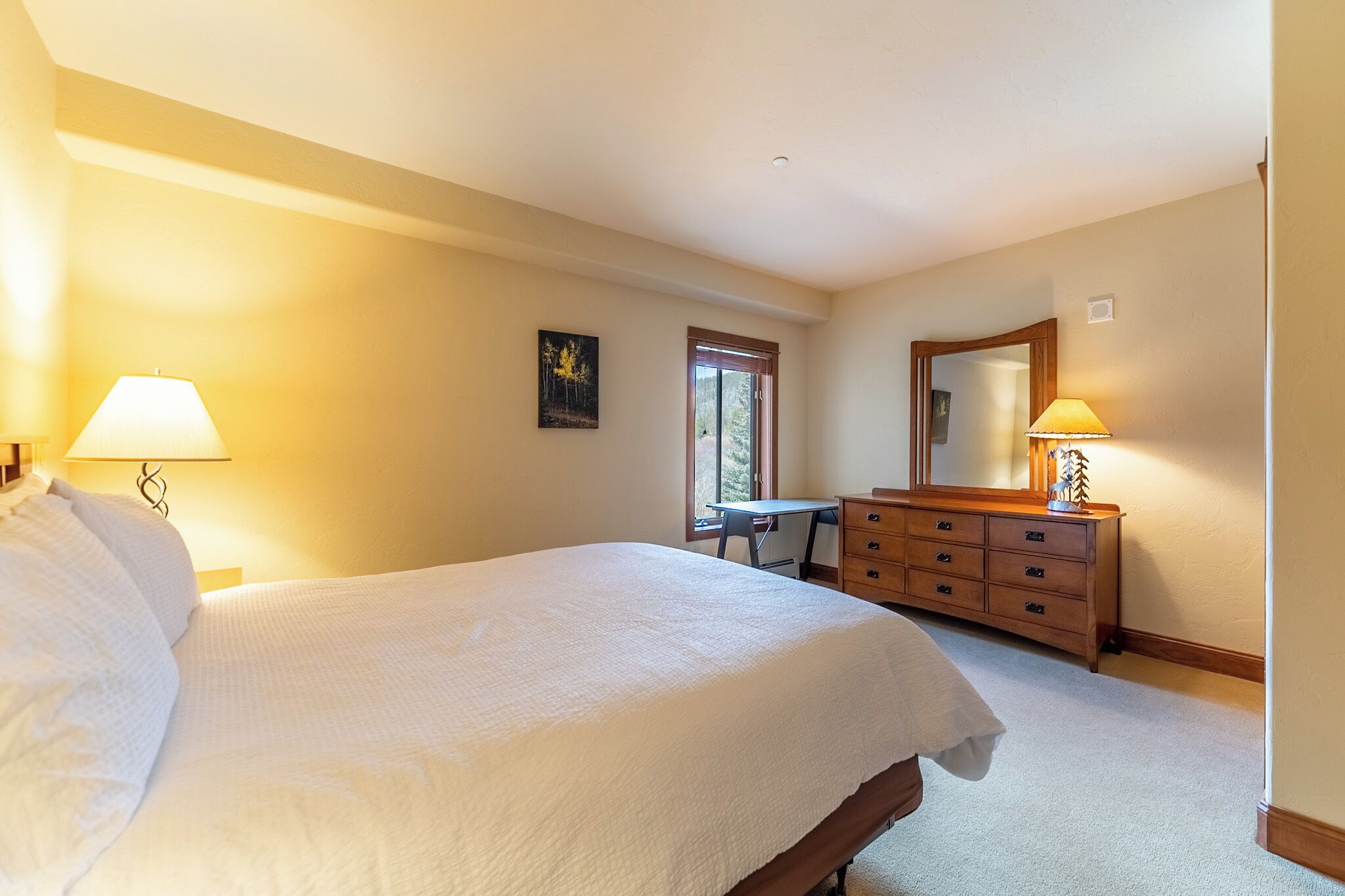 A bedroom with a large bed, a desk, a dresser, and a window with a view of trees.