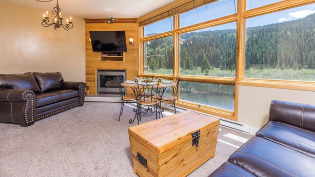 Ein gemütliches Wohnzimmer mit einer Ledersofa, einem Holztisch und einem Esstisch mit Stühlen. Ein großes Fenster bietet Blick auf eine bewaldete Gegend.