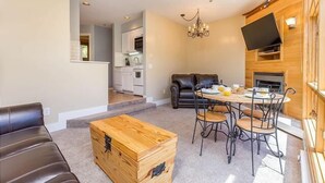 Ein gemütliches Wohnzimmer mit einer Couch, einer Holzkiste und einem Esstisch mit Stühlen. Im Hintergrund ist eine Küche zu sehen.