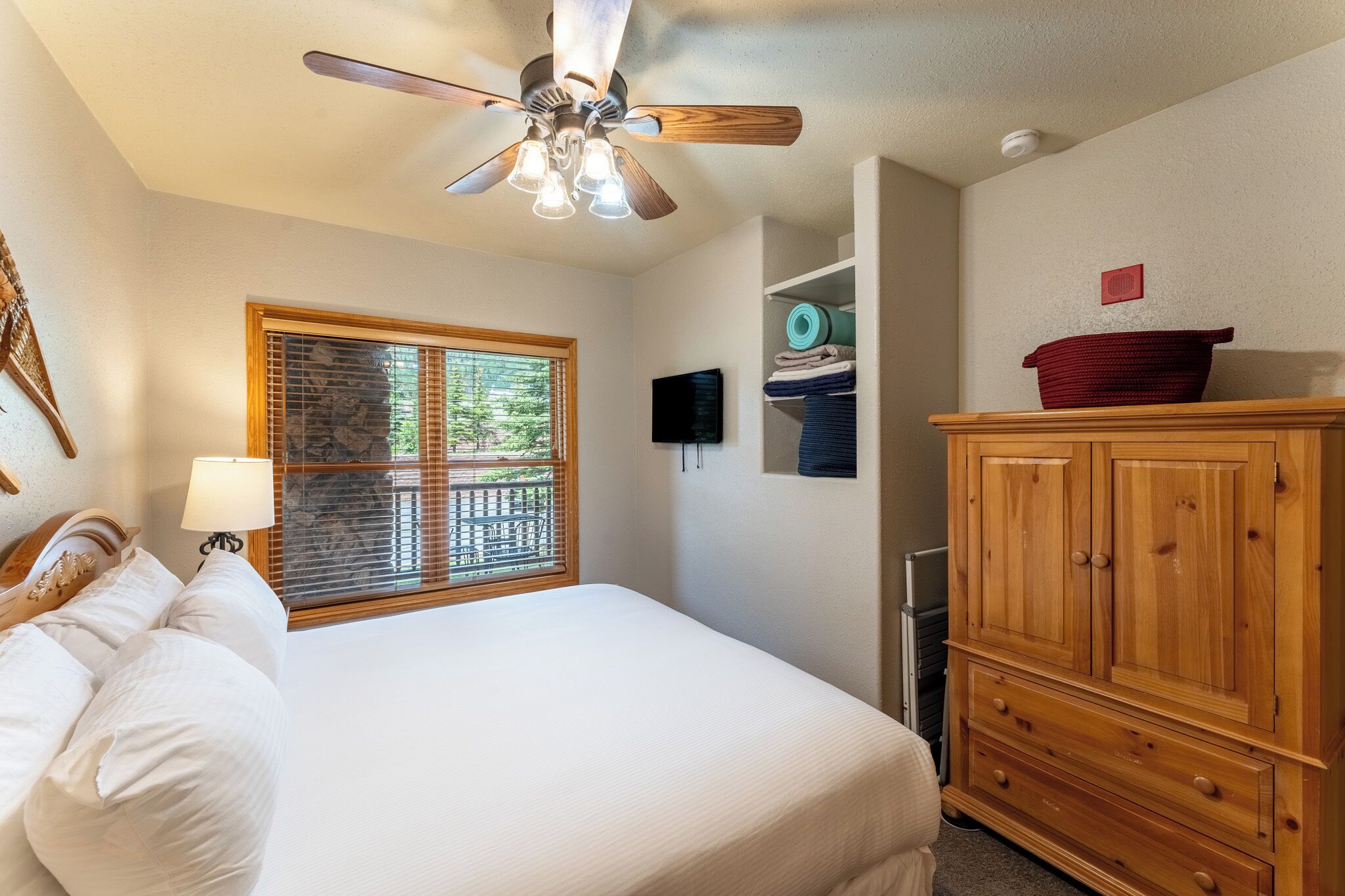 A bedroom with a bed, a ceiling fan, a window with blinds, a TV, and a wooden cabinet.