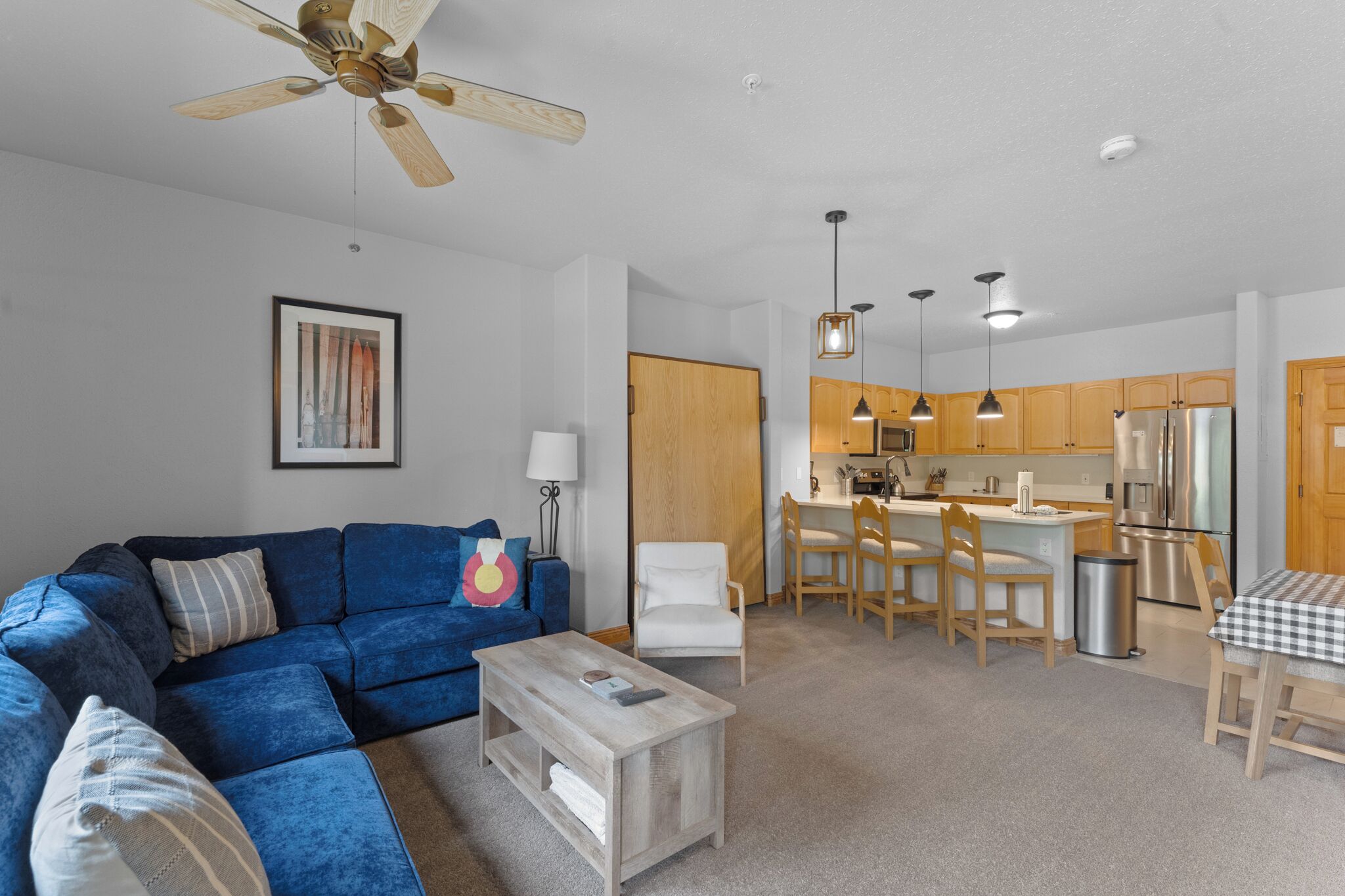A modern living room with a blue sofa, a wooden coffee table, and a ceiling fan. Adjacent is a kitchen with wooden cabinets, stainless steel appliances, and pendant lighting.