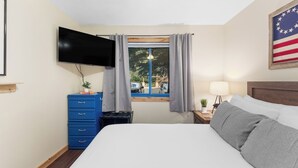 Hotellihuone, jossa on sänky, seinälle asennettu televisio, verhoilla varustettu ikkuna, yöpöytä lampun kanssa ja pieni pöytä, jossa on kasvi.