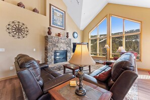 A cozy living room with leather sofas, a stone fireplace, and large windows offering a mountain view.