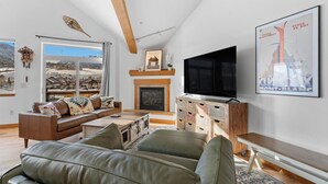 A modern living room with a fireplace, leather sofas, a television, and a large window offering a mountain view. - Retreat on the Blue, Unit E2 : 150 (Silverthorne)