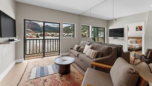 A modern living room with a large sofa, a coffee table, and a TV. There are large windows offering a view of mountains and a building. - Greens at Copper #215 (Copper Mountain)