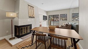A modern living room with a fireplace, a dining table with chairs, a sofa, and a large window with a view. - Greens at Copper #215 (Copper Mountain)