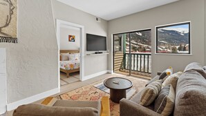 A living room with a sofa, a coffee table, and a TV. There is a balcony with a view of mountains. - Greens at Copper #215 (Copper Mountain)