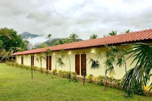 A row of yellow buildings with red roofs, surrounded by grass and palm trees, under a cloudy sky. - The Creek Garden Resort (Mae Hong Son)