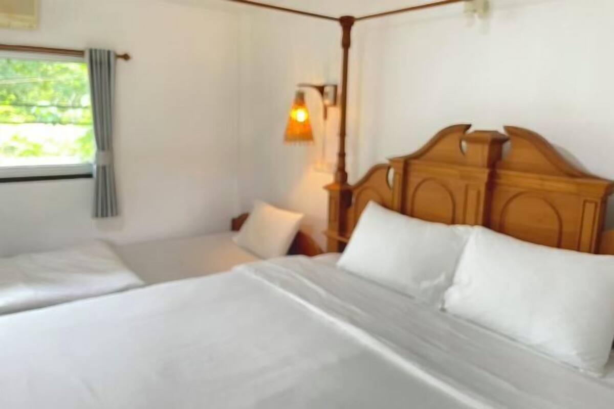 A room with a large bed, a smaller bed, a wooden headboard, a window with curtains, and a wall-mounted lamp.