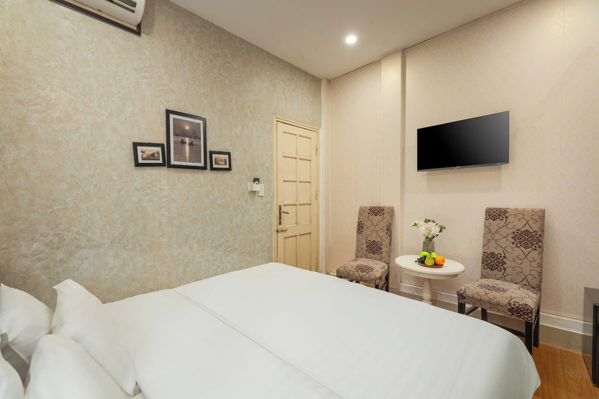 A hotel room with a large bed, two chairs, a small table with a vase of flowers, a flat-screen TV mounted on the wall, and a door.