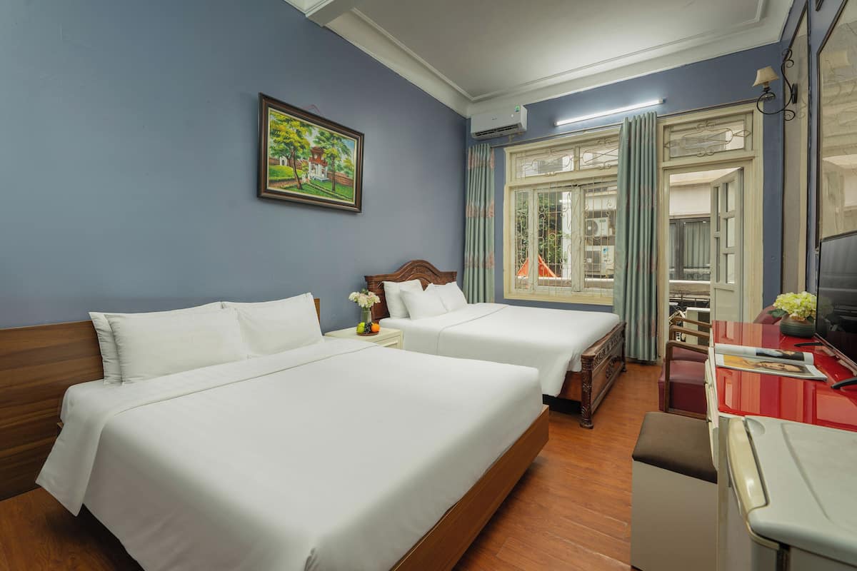 A hotel room with two beds, a wooden headboard, a small table with a vase, and a window with curtains.