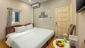 A neatly made bed in a hotel room with a wooden headboard, a small table with fruit and a book, and a door with a decorative panel. - Hanoi Old quarter Central Hotel (Hanoi)