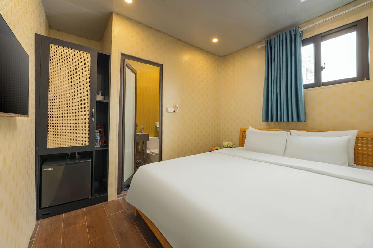 A hotel room with a bed, a small refrigerator, a mirror, a window with curtains, and a bathroom visible through an open door.