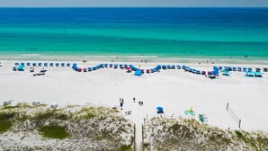 Une plage de sable blanc bordée par une mer d’un bleu limpide. Des parasols bleus et des chaises de plage sont disposés en demi-cercle.