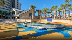 Un espace ludique pour enfants complexe, avec une surface de jeu colorée, entouré de palmiers et d’un bâtiment doté de balcons.