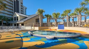 Uma área infantil de piscina complexa, com revestimento colorido, cercada por palmeiras e um edifício com varandas.