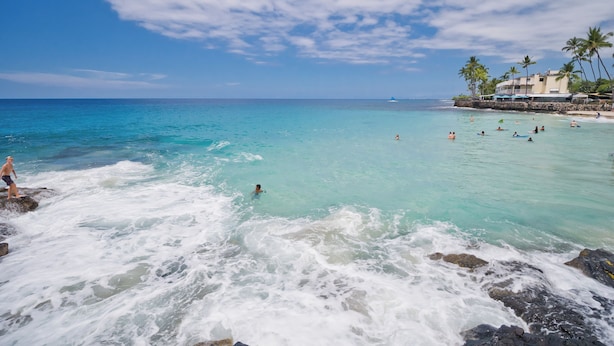 Una playa con agua turquesa cristalina, gente nadando y parada sobre rocas, y un edificio con palmeras al fondo.