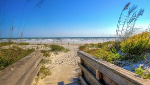 A wooden boardwalk leading to a beach with sand and sparse vegetation. - Beachfront Bliss by the Surf | Inlet #605 (New Smyrna Beach)