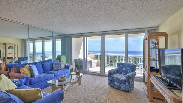 A spacious living room with a large glass door leading to a balcony with a view of the ocean. There is a blue sofa, a patterned armchair, and a television on a wooden stand. - Beachfront Bliss by the Surf | Inlet #605 (New Smyrna Beach)