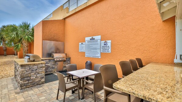 An outdoor dining area with stone walls, a grill, and seating. - Beachfront Bliss by the Surf | Inlet #605 (New Smyrna Beach)