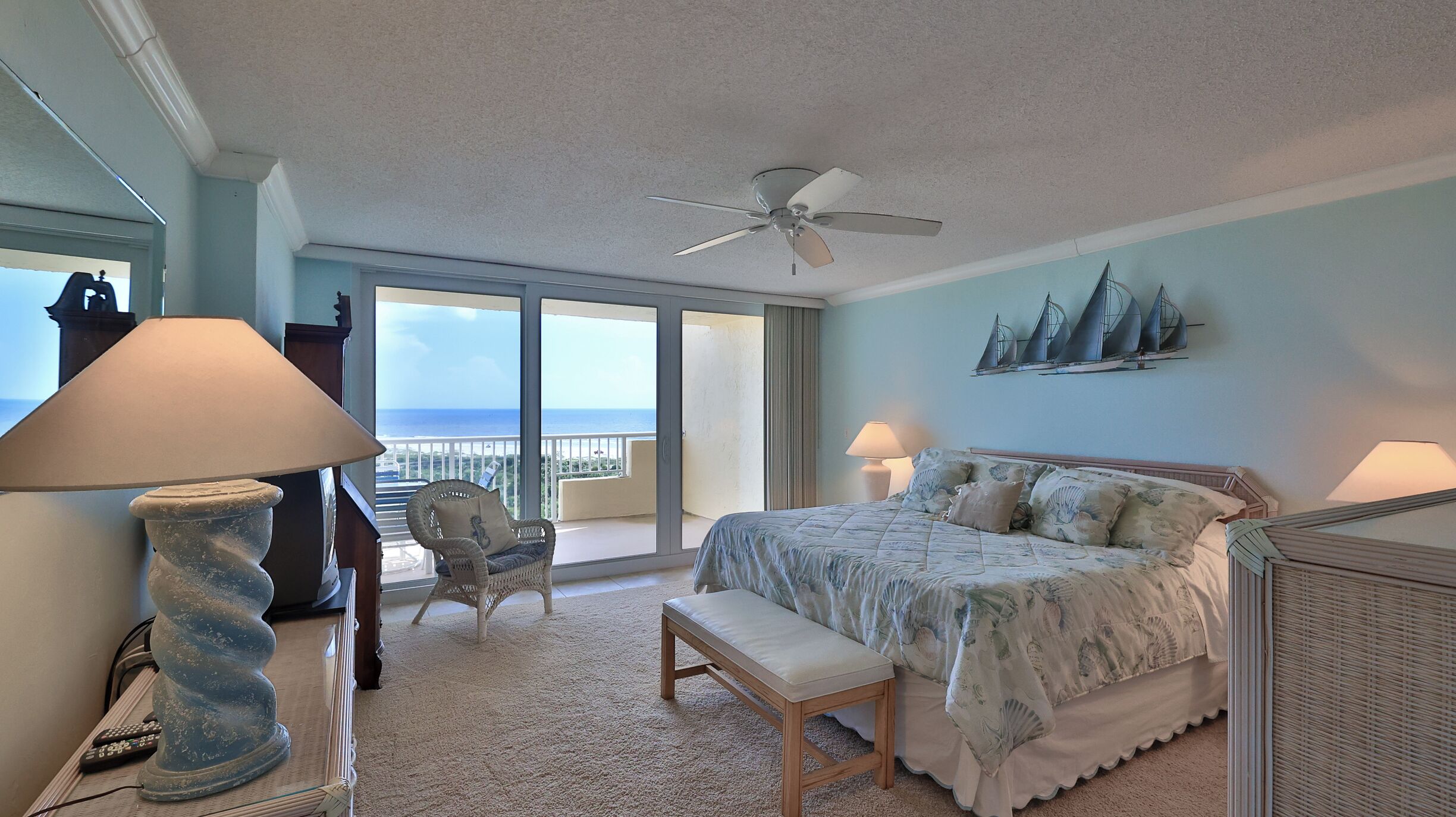 A bedroom with a bed, a bench, a chair, a ceiling fan, and a view of the ocean.