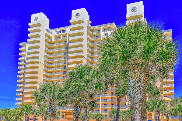 Un hôtel à plusieurs étages doté de balcons, avec des palmiers au premier plan et un ciel bleu dégagé.