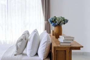 Una cama con ropa blanca y almohadas, una mesita de noche de madera con un florero y libros, y una ventana con cortinas translúcidas.