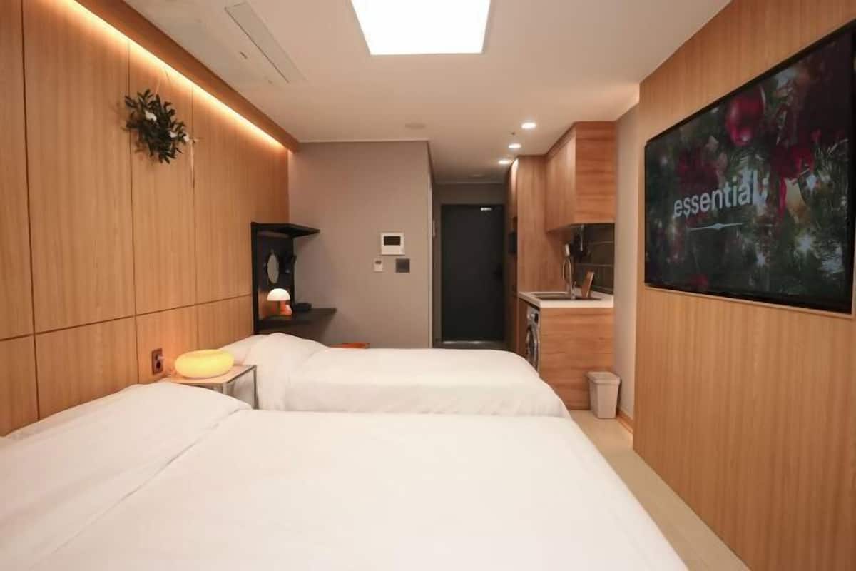 A hotel room with two beds, a small desk, a nightstand, a wall-mounted TV, and a bathroom visible through an open door.