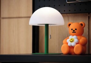 Eine Lampe mit weißem Schirm und grünem Fuß, ein Teddybär mit lächelndem Gesicht und ein Holzuntergrund.