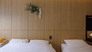 Quarto de hotel com cabeceira de madeira, duas camas, mesa de cabeceira com abajur e planta fixada na parede.