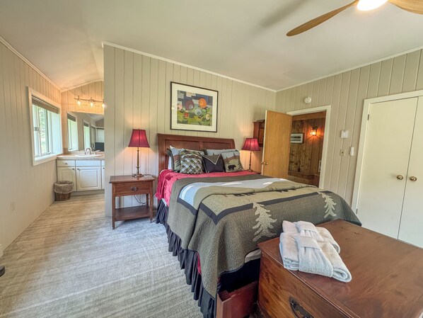 Un dormitorio con cama, lámparas de noche, cabecera de madera, un cuadro, una puerta y un pequeño cofre de madera.