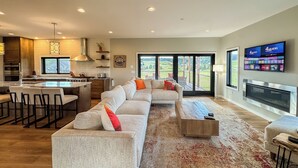 A modern living room with a large sectional sofa, a dining area with bar stools, a kitchen with a central island, and a fireplace.