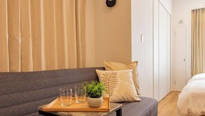 A modern hotel room with a grey sofa, a wooden side table with glasses and a plant, and a bed with white bedding. - Tokyo Asakusa House (Tokyo)