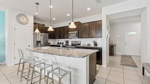 A modern kitchen with dark wood cabinets, a granite countertop, stainless steel appliances, and a central island with bar stools. - Spacious 5BR Villa w/ Private Pool – near Disney! (Kissimmee)
