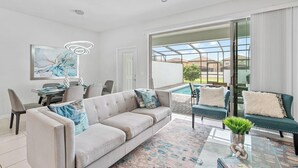 A modern living room with a sofa, chairs, a dining table, and a glass-enclosed patio area with a pool. - Spacious 5BR Villa w/ Private Pool – near Disney! (Kissimmee)